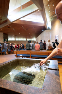 A hand reaches towards a baptismal font in a modern church with wooden ceilings and skylights. People gathered in the background near pews and a stone wall.