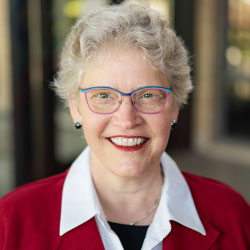 Robin Steinke is wearing a red blazer and white collared shirt. She has short wavy hair and blue and pink glasses.