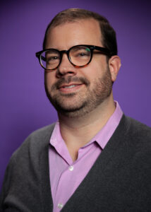 Cody Sanders has short hair, dark-framed glasses, and a light beard. He is wearing a purple collared shirt and gray cardigan.