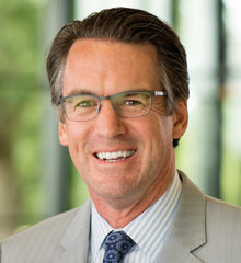 Matt Skinner is wearing a gray suit, striped shirt with blue tie, and glasses. He has short dark hair and is smiling at the camera.