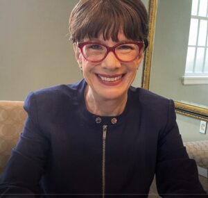 Amy-Jill Levine is wearing a navy zippered blazer and a pair of red glasses.