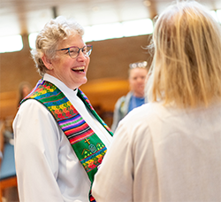 A person in a white robe and colorful stole smiling at another person in a church setting.
