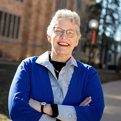 A person with short gray hair and glasses smiles with arms crossed, standing outdoors in front of a brick building and trees. They are wearing a blue jacket over a light shirt.