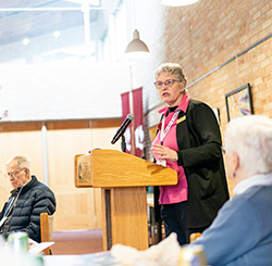 A woman speaking at a podium in a well-lit room with a brick wall background and seated attendees.