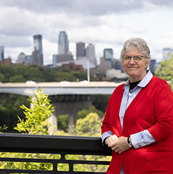 Person in a red cardigan standing by a railing with a city skyline and bridge in the background.
