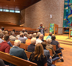 A person speaks to an audience in a church with brick walls and colorful stained-glass windows.