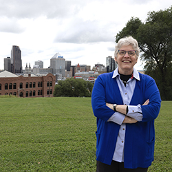 A woman with short white hair and glasses, wearing a blue cardigan over a button-down shirt, standing with arms crossed in a grassy field with a city skyline in the background.