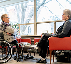 Robin Steinke and Rolf Jacobson seated in red armchairs having a conversation near a large window.