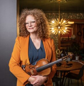 Publicity photo of songwriter Becky Buller holding a fiddle and standing in the entryway to dining-living room space
