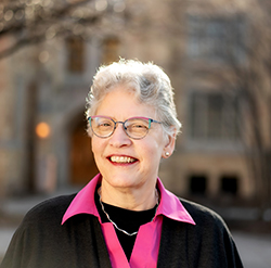 A smiling woman with short, curly grey hair and glasses wearing a pink collared shirt under a dark sweater, with a blurred stone building in the background.