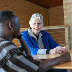 A smiling woman with short gray hair and glasses, wearing a blue jacket, sits at a table engaged in conversation with a man whose back is to the camera.