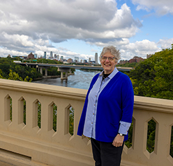 A smiling woman with short gray hair wearing a light blue shirt and a royal blue cardigan, standing outdoors on a bridge with a river and city skyline in the background.