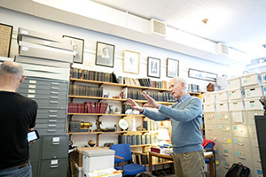 Two people in a library or office, surrounded by shelves filled with books and files.