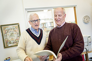 Two older men reading an open book together indoors.