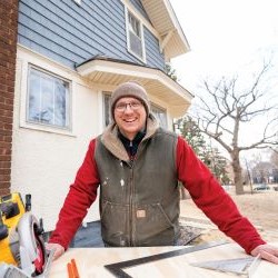 Person in winter clothing smiling at a workbench in front of a house.