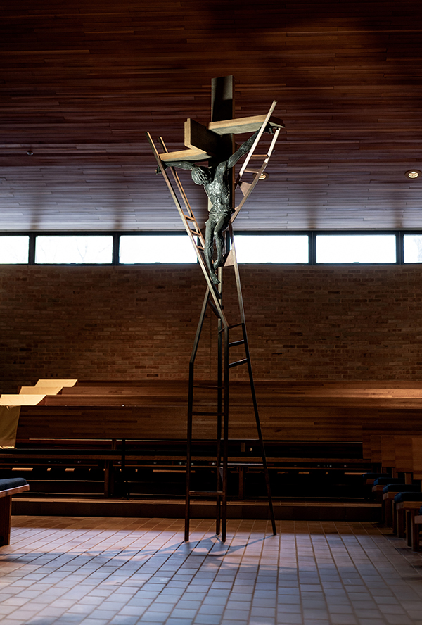 Modern crucifix sculpture standing on tall metal framework in a spacious room with wooden ceiling and brick walls.