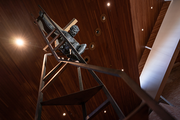 A sculpture of a human figure climbing a tall metal ladder against a wooden ceiling with lights.