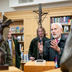 A man in a library explaining an abstract metal sculpture to a group of people.