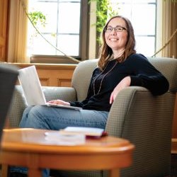 Person sitting in an armchair with a laptop, smiling in a warmly lit room.