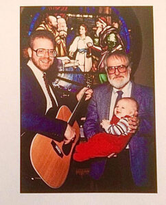 Two men, one with a guitar, and a baby in front of a stained glass window.