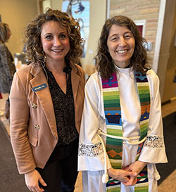 Two women smiling indoors, one wearing a beige blazer and the other in religious attire with a colorful stole.
