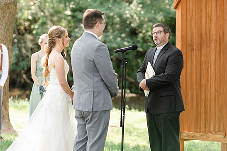 An outdoor wedding ceremony with a bride, groom, and officiant standing next to a wooden structure.
