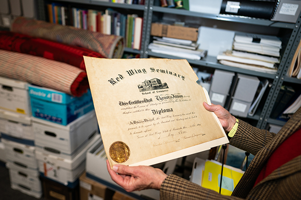 Person holding a diploma from Red Wing Seminary in a storage room with shelves of books and boxes.