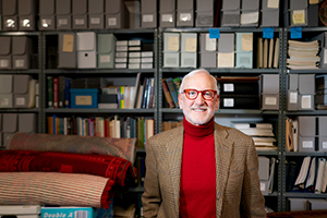 A man in a brown blazer and red turtleneck stands in front of shelves filled with boxes and books.