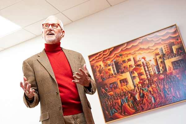 An older man in a brown blazer and red turtleneck gestures while standing in front of a colorful urban painting.