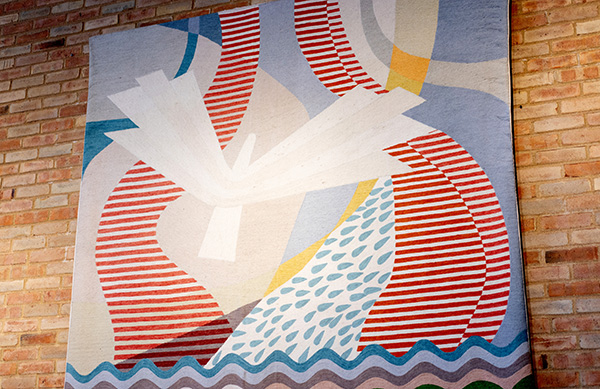 Abstract textile art on a brick wall with red and white stripes, teardrop shapes, and wave patterns in blue and lavender tones.
