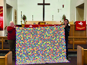 Two people holding a colorful quilt inside a church with an altar and cross in the background.