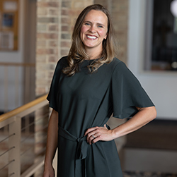 A woman in a stylish dark green dress stands confidently near a staircase, with a modern interior featuring brick walls in the background.