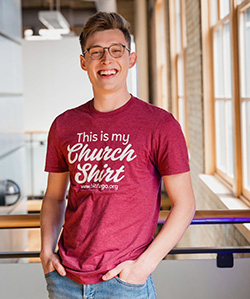 A smiling young man in glasses wears a maroon t-shirt that reads, "This is my Church Shirt"