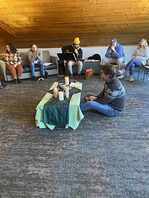 A small group of people sit around a low table with candles, with one person playing a guitar, in a casual worship or discussion setting.