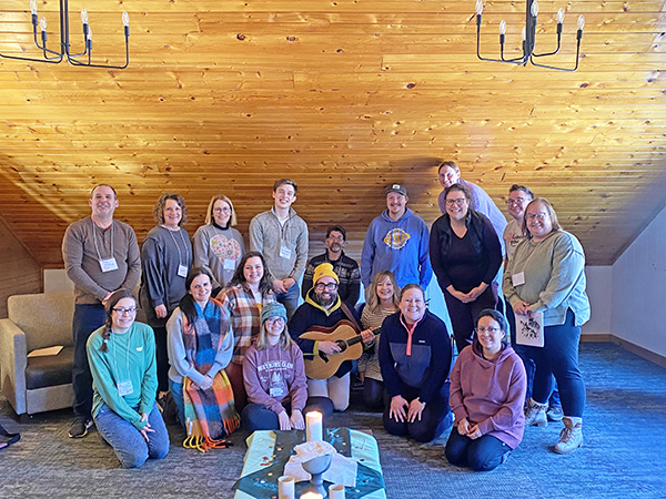 A group of approximately 18 adults pose for a group photo in a room with wood-paneled walls and ceiling, with one person in the center holding a guitar.
