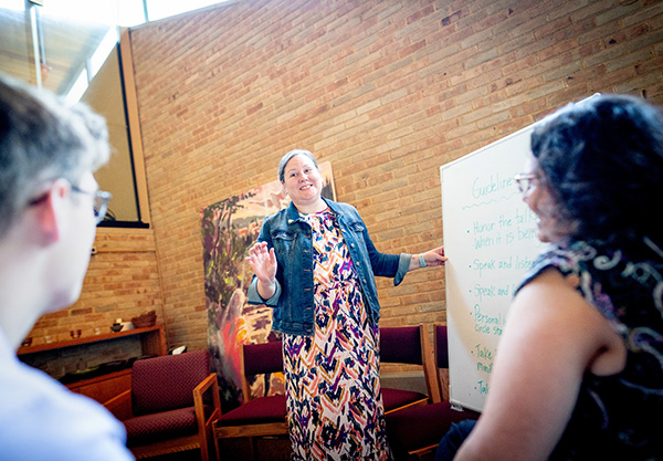 A woman in a floral dress and denim jacket points to a whiteboard listing "Guidelines" while speaking to two people in a brick-walled room.