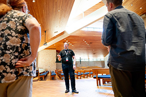 Three people talking inside a modern chapel with wood paneling and blue pews.