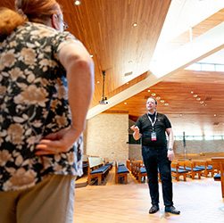 Two people talking inside a modern chapel with wood paneling and blue pews.