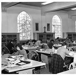 Black and white photo of a group of students studying at large tables in a library with tall arched windows and religious imagery on the back wall.