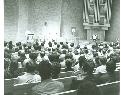 A spacious, modern chapel interior with a large circular rug, a stone altar, and many chairs arranged in a wide semicircle.