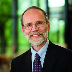 Headshot of a smiling man with a beard, wearing glasses, a suit jacket, and a tie against a soft-focus background.