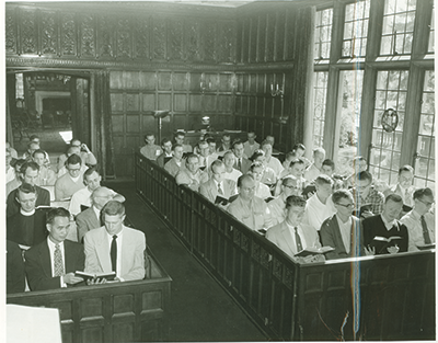 A traditional chapel setting with a central wooden altar, a large cross against a brick wall, and rows of chairs facing forward.