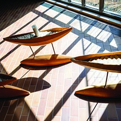 Wooden canoe-shaped sculptures casting shadows on a brick floor inside a sunlit room.