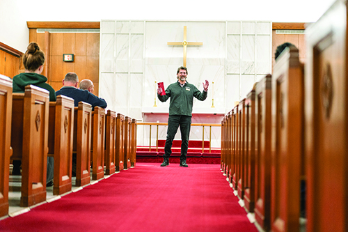 A presenter standing on a stage in a large hall, gesturing toward a PowerPoint slide projected on a screen for an audience.