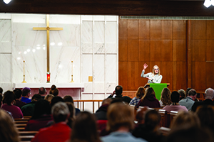 A wide shot of an audience seated at tables, listening to a presentation in a large hall with wood paneling and decorative ceiling trusses.