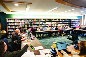 A large group of people sit around a U-shaped table with laptops in a library room lined with bookshelves.