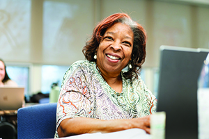 Close-up of a smiling woman sitting with a laptop at a table in a brightly lit seminar room.