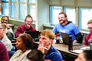 A smiling man in a blue Luther Seminary t-shirt sits with a laptop at a long table among other students in a classroom.