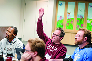 A man in a maroon Luther Seminary sweatshirt raises his hand to ask a question during a lecture or seminar.