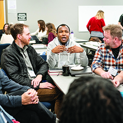 A man in a grey hoodie gestures with his hands while speaking to a small group gathered around a classroom table.